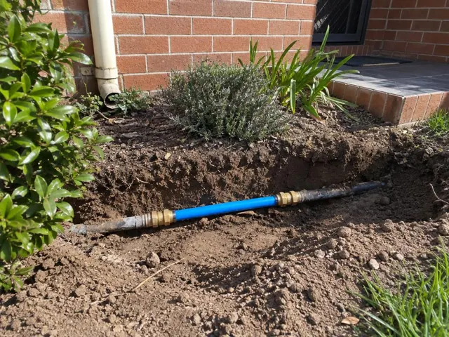 Exposed repaired water pipe in a soil trench beside a brick house and garden bed in Newcastle